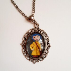 Pu&ograve; includere: Una collana con catena d'argento con un ciondolo ovale che presenta una ragazza dei cartoni animati con i capelli blu che indossa un impermeabile giallo. Il ciondolo ha una cornice d'argento decorativa.