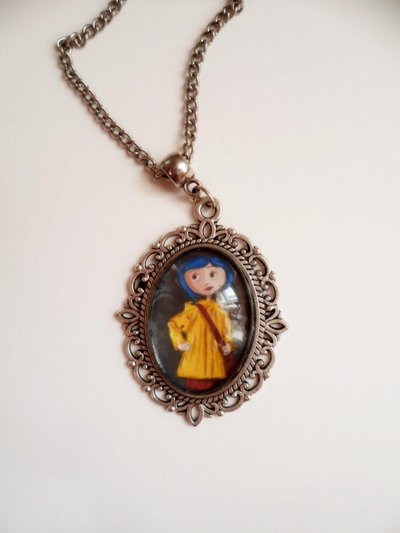 Pu&ograve; includere: Una collana con catena d'argento con un ciondolo ovale che presenta una ragazza dei cartoni animati con i capelli blu che indossa un impermeabile giallo. Il ciondolo ha una cornice d'argento decorativa.