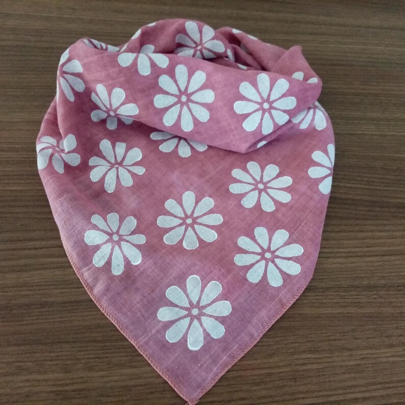 May include: A dusty rose bandana with a white daisy pattern. The square scarf is folded and arranged on a wooden surface. The floral design is evenly spaced across the fabric, creating a simple, yet elegant accessory.
