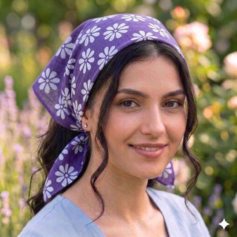 May include: A purple headscarf with a white floral pattern. The head covering is tied at the back and worn on the head. The background is a garden with flowers.