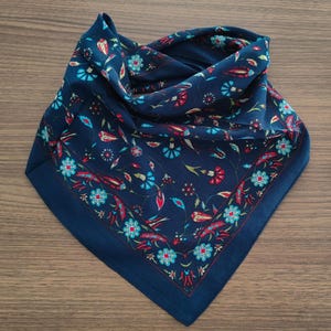 May include: A square navy blue bandana featuring a repeating floral pattern in shades of turquoise, red, and white. The fabric displays stylized flowers and leaves arranged in a symmetrical design with a distinct border framing the central motif.