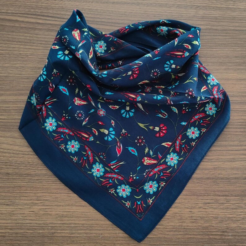 May include: A square navy blue bandana featuring a repeating floral pattern in shades of turquoise, red, and white. The fabric displays stylized flowers and leaves arranged in a symmetrical design with a distinct border framing the central motif.