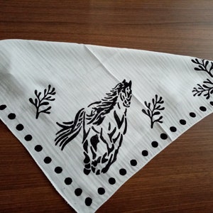 K&ouml;nnte beinhalten: Ein wei&szlig;es, dreieckiges Bandana mit einem schwarzen Siebdruckmotiv eines galoppierenden Pferdes in der Mitte. Der Stoff hat eine feine vertikale Streifenstruktur und ist mit einer Reihe schwarzer Punkte und stilisierter Blattmotive verziert.