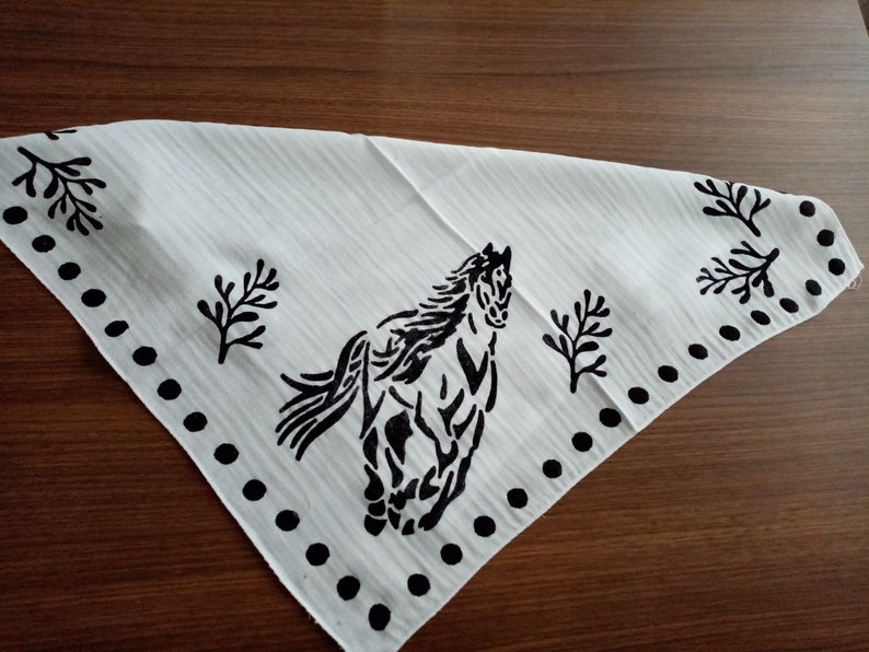 K&ouml;nnte beinhalten: Ein wei&szlig;es, dreieckiges Bandana mit einem schwarzen Siebdruckmotiv eines galoppierenden Pferdes in der Mitte. Der Stoff hat eine feine vertikale Streifenstruktur und ist mit einer Reihe schwarzer Punkte und stilisierter Blattmotive verziert.