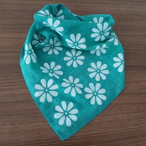 May include: A folded teal bandana with a white daisy pattern. The bandana is square-shaped and made of a soft fabric. The daisy pattern is evenly spaced across the fabric. The bandana is likely used as a fashion accessory.