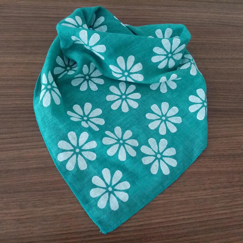 May include: A folded teal bandana with a white daisy pattern. The bandana is square-shaped and made of a soft fabric. The daisy pattern is evenly spaced across the fabric. The bandana is likely used as a fashion accessory.