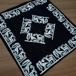 May include: Square black textile with white decorative patterns. The border features a repeating design of stylized animals and foliage. The center has four smaller, similar motifs. The textile is likely a decorative item, such as a scarf or a small tablecloth.
