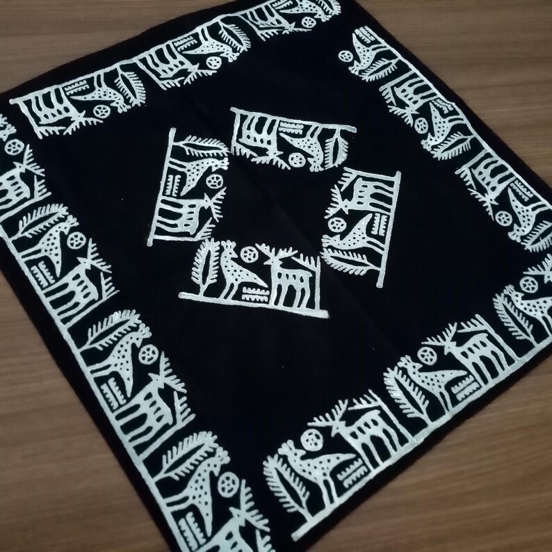 May include: Square black textile with white decorative patterns. The border features a repeating design of stylized animals and foliage. The center has four smaller, similar motifs. The textile is likely a decorative item, such as a scarf or a small tablecloth.
