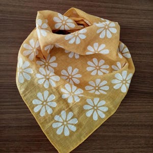 May include: Mustard yellow bandana with a white daisy pattern. The square scarf is folded and arranged to show the floral design. The fabric is soft and lightweight, suitable for various uses.