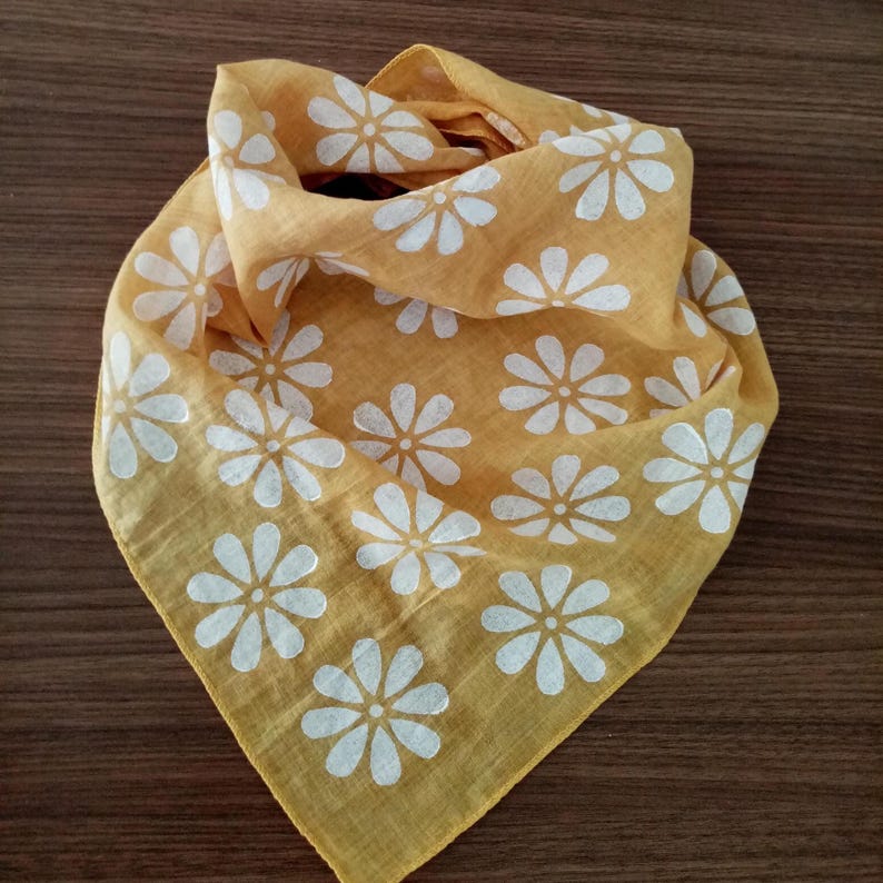 May include: Mustard yellow bandana with a white daisy pattern. The square scarf is folded and arranged to show the floral design. The fabric is soft and lightweight, suitable for various uses.