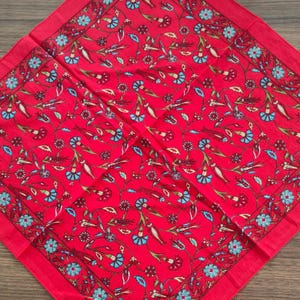 May include: A square fabric scarf featuring a vibrant red background with a repeating pattern of stylized flowers and vines in shades of light blue, dark red, and cream. The design includes a decorative border with small blue floral motifs.