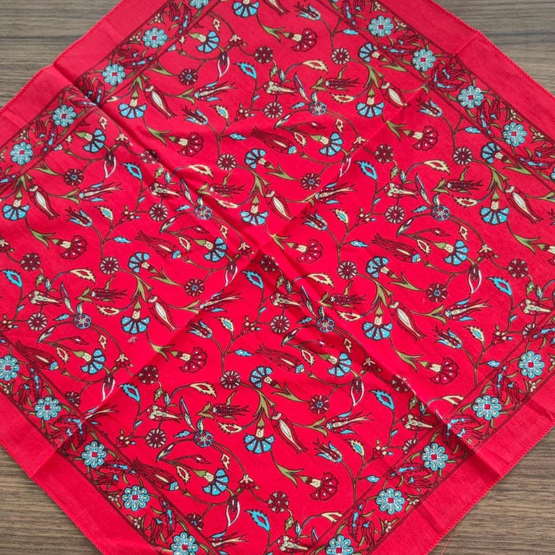 May include: A square fabric scarf featuring a vibrant red background with a repeating pattern of stylized flowers and vines in shades of light blue, dark red, and cream. The design includes a decorative border with small blue floral motifs.