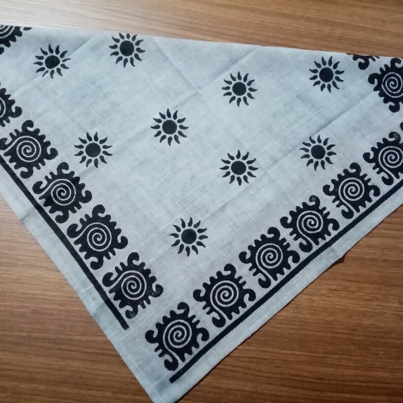 May include: A light gray bandana with a repeating pattern of black suns and a black border with a spiral design. The bandana is folded in a triangle shape and is made of a soft fabric.