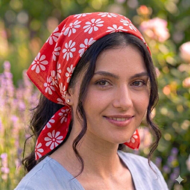 May include: A red headscarf with a white daisy pattern. The head covering is tied at the back of the head. The background is a garden with flowers.