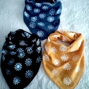 May include: Three bandanas in navy blue, black, and mustard yellow. Each bandana has a repeating pattern of white suns. The bandanas are folded and arranged on a white surface.