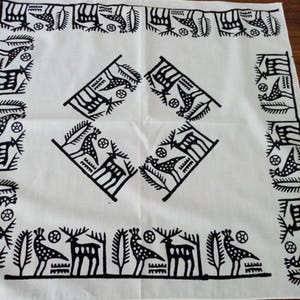 May include: White square textile with a repeating black pattern. The design features stylized deer, birds, and geometric shapes. The border has a repeating pattern of birds and other shapes. The center has four diamond-shaped designs.