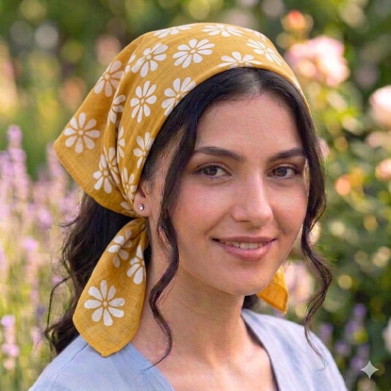 May include: Mustard yellow headscarf with a white daisy pattern. The head covering is tied at the back and worn on the head. The background is a garden with pink and purple flowers. The person is wearing a light blue shirt.