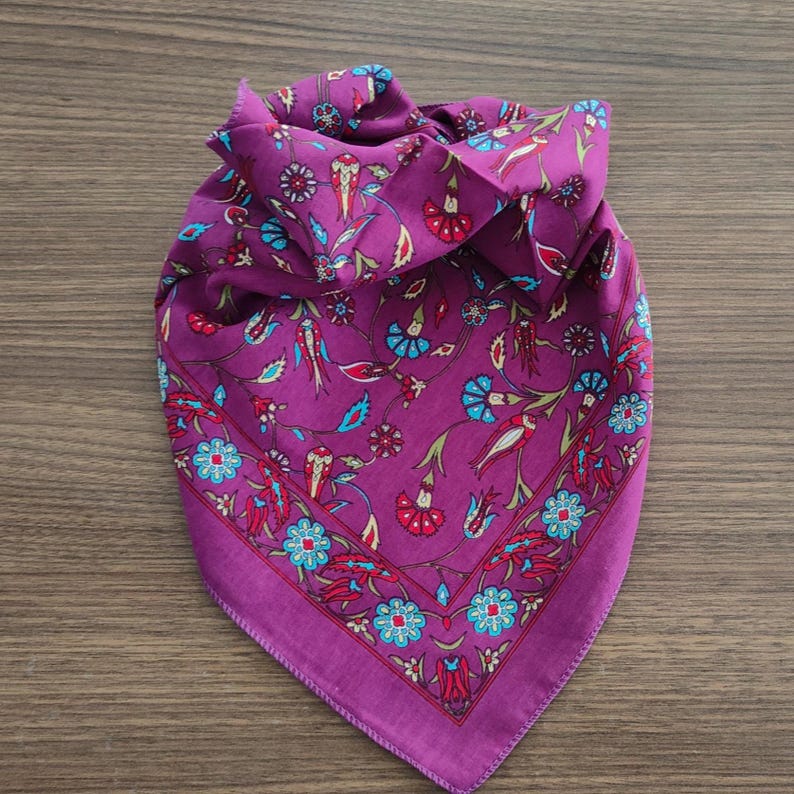 May include: A square purple scarf featuring a repeating pattern of stylized flowers and birds in shades of red, light blue, and green. The design includes a distinct floral border that frames the central motif, creating a cohesive look.