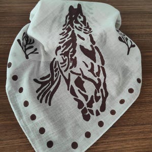 K&ouml;nnte beinhalten: Ein dreieckiges, cremewei&szlig;es Bandana mit einem dunkelbraunen Siebdruckmotiv eines Pferdekopfes in der Mitte. Das Design enth&auml;lt kleine dunkelbraune Punkte entlang der unteren Kanten sowie stilisierte Zweigmotive an den oberen Seiten.