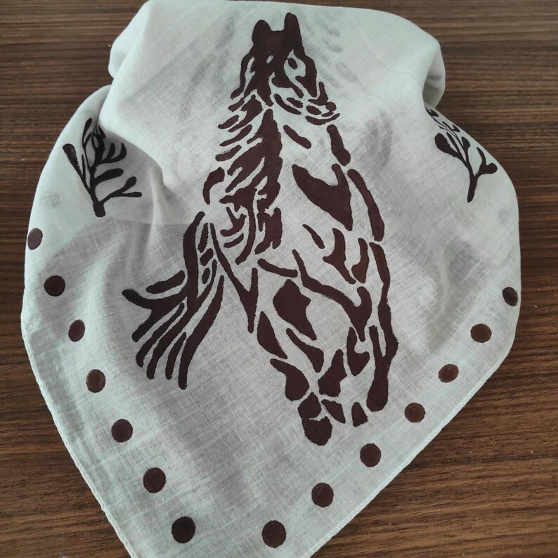 K&ouml;nnte beinhalten: Ein dreieckiges, cremewei&szlig;es Bandana mit einem dunkelbraunen Siebdruckmotiv eines Pferdekopfes in der Mitte. Das Design enth&auml;lt kleine dunkelbraune Punkte entlang der unteren Kanten sowie stilisierte Zweigmotive an den oberen Seiten.