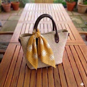 May include: A beige tote bag with brown leather handles and accents, and a mustard yellow scarf with white sun designs tied to the handle. The bag is on a wooden table.
