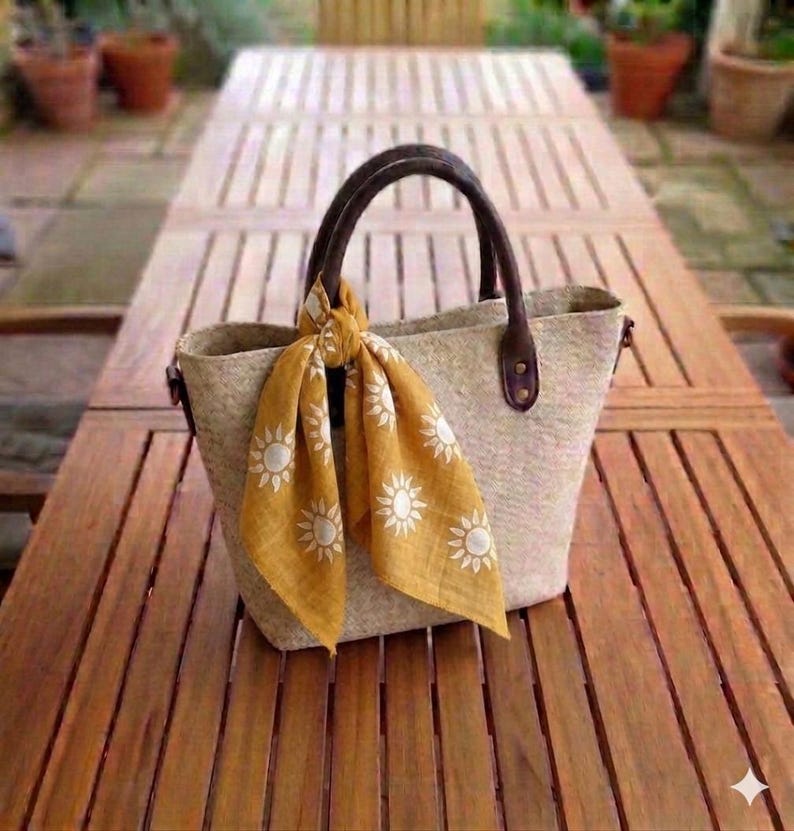 May include: A beige tote bag with brown leather handles and accents, and a mustard yellow scarf with white sun designs tied to the handle. The bag is on a wooden table.