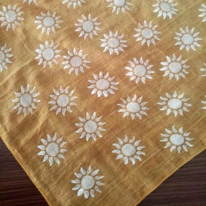 May include: Mustard yellow fabric featuring a repeating pattern of white suns. The material has a slightly textured appearance and is folded, showing a corner. The suns are stylized with a central circle and radiating lines.