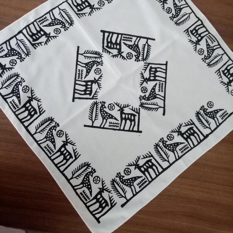May include: White square cloth with a black geometric pattern. The design features stylized birds, deer, and abstract shapes. The border has a repeating pattern of birds and deer. The cloth is folded diagonally, showing the design in the center and along the edges.