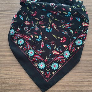 May include: A black square scarf featuring a vibrant floral pattern with red, turquoise, and green botanical motifs. The design includes a distinct border of repeating blue flowers and red tulip-like shapes, set against a solid black background.
