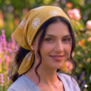 May include: Mustard yellow headscarf featuring white sunburst patterns. The head covering is tied at the back. The background is a blurred garden with pink and purple flowers.