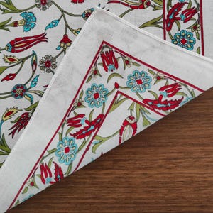 May include: A white fabric textile featuring a repeating floral pattern with red tulips, light blue blossoms, and green stems. The design includes a distinct border with a red line framing the floral motifs, creating a structured visual layout.