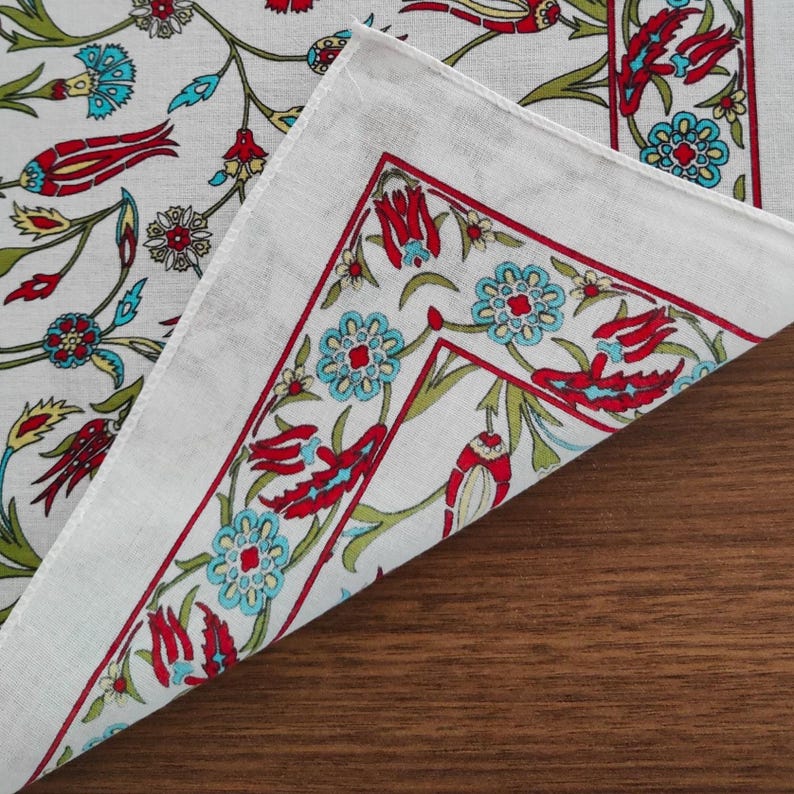 May include: A white fabric textile featuring a repeating floral pattern with red tulips, light blue blossoms, and green stems. The design includes a distinct border with a red line framing the floral motifs, creating a structured visual layout.