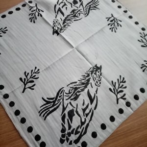 K&ouml;nnte beinhalten: Ein quadratisches wei&szlig;es Stoffbandana mit einem schwarzen Siebdruckmotiv von zwei galoppierenden Pferden. Der Rand ist mit einer Reihe schwarzer Punkte verziert und der Hintergrund ist mit kleinen schwarzen Zweigmotiven zwischen den Pferden akzentuiert.
