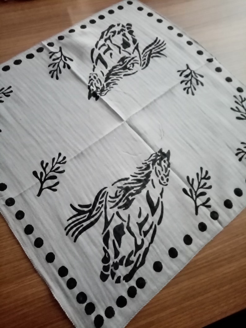 K&ouml;nnte beinhalten: Ein quadratisches wei&szlig;es Stoffbandana mit einem schwarzen Siebdruckmotiv von zwei galoppierenden Pferden. Der Rand ist mit einer Reihe schwarzer Punkte verziert und der Hintergrund ist mit kleinen schwarzen Zweigmotiven zwischen den Pferden akzentuiert.