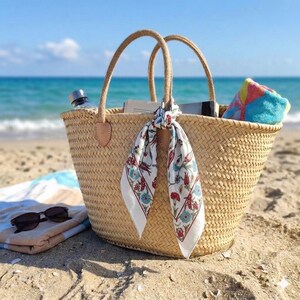 May include: A woven straw tote bag with two handles, featuring a white silk scarf tied around one handle with a colorful floral pattern. The bag contains a book and a folded multi-colored towel, with a pair of sunglasses resting nearby.