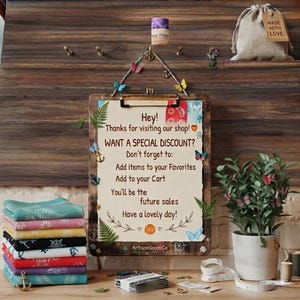 May include: A wooden clipboard with a cream-colored sign featuring text about shop discounts, decorated with small butterfly and anchor accents. A stack of folded colorful patterned fabric squares sits to the left, and a green plant is in a pot.