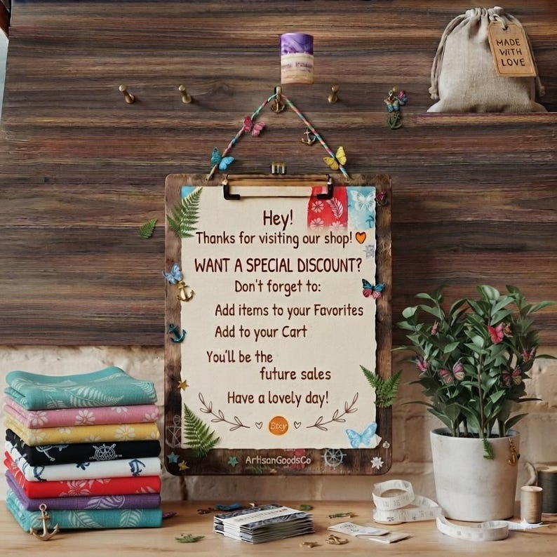 May include: A wooden clipboard with a cream-colored sign featuring text about shop discounts, decorated with small butterfly and anchor accents. A stack of folded colorful patterned fabric squares sits to the left, and a green plant is in a pot.
