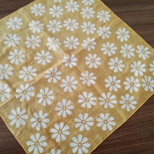 May include: A square, yellow handkerchief with a repeating pattern of white daisy flowers. The fabric is folded diagonally, showcasing the floral design. The handkerchief is made of a soft, lightweight material.