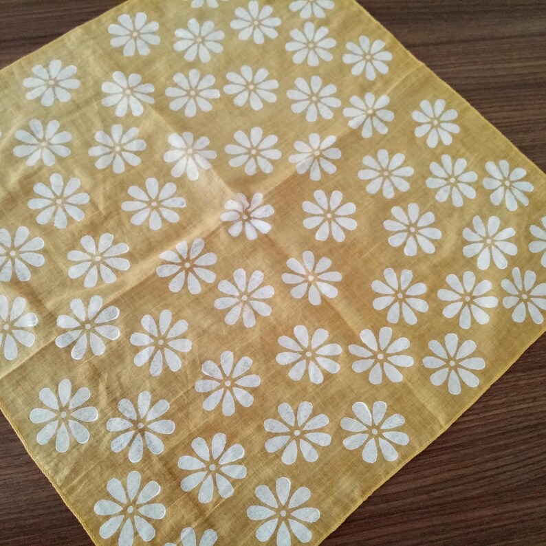 May include: A square, yellow handkerchief with a repeating pattern of white daisy flowers. The fabric is folded diagonally, showcasing the floral design. The handkerchief is made of a soft, lightweight material.