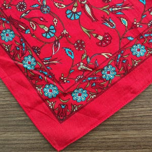 May include: A square fabric bandana featuring a vibrant red background with a repeating floral pattern in shades of bright blue, cream, and dark red. The design includes a decorative border framing the central motif of stylized flowers and vines.
