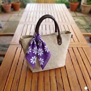 May include: A beige tote bag with brown leather handles and accents. A purple scarf with white daisy patterns is tied to the handle. The bag is sitting on a wooden table.