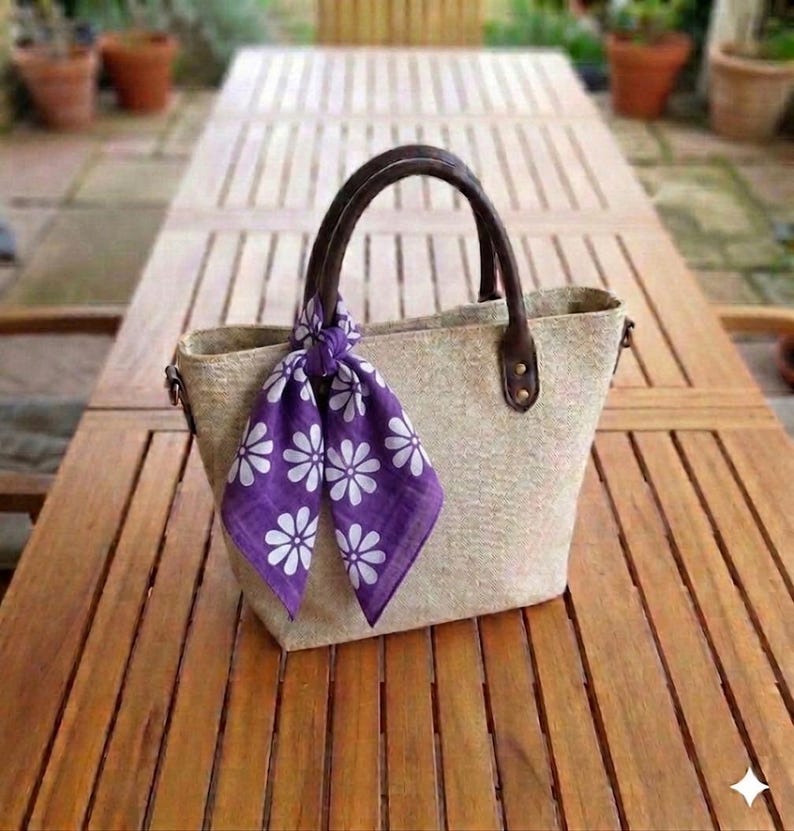 May include: A beige tote bag with brown leather handles and accents. A purple scarf with white daisy patterns is tied to the handle. The bag is sitting on a wooden table.