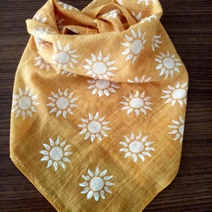 May include: Mustard yellow bandana featuring a repeating pattern of white suns. The bandana is folded, suggesting a lightweight fabric. The suns have a circular centre with radiating lines.