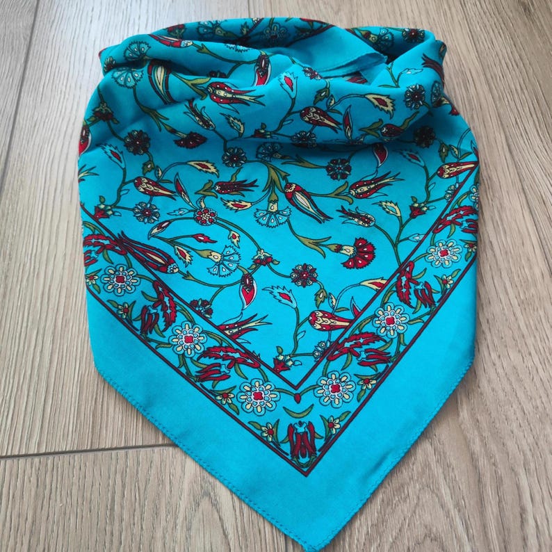 May include: A square scarf in a vibrant turquoise blue color featuring a detailed floral and botanical pattern. The design includes stylized red, white, and green flowers and leaves with dark brown outlines, arranged in a repeating motif.