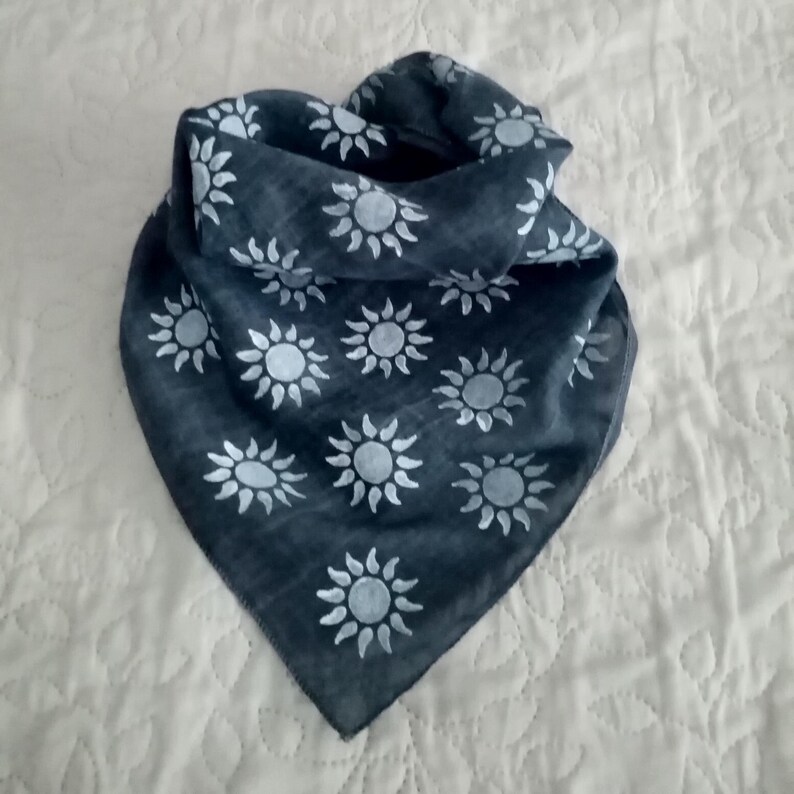 May include: A dark grey bandana scarf featuring a repeating pattern of white sunburst designs. The scarf is folded and draped, highlighting the fabric's texture and the contrast between the dark background and the bright white suns. The scarf is likely made of a lightweight material.