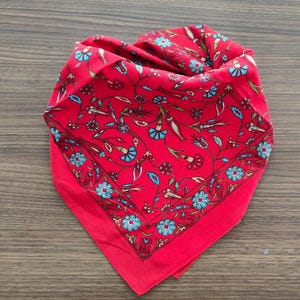 May include: A square bandana featuring a vibrant red background with a repeating floral pattern in shades of light blue, dark red, and green. The design includes stylized flowers and leaves arranged in a symmetrical layout with a decorative border.