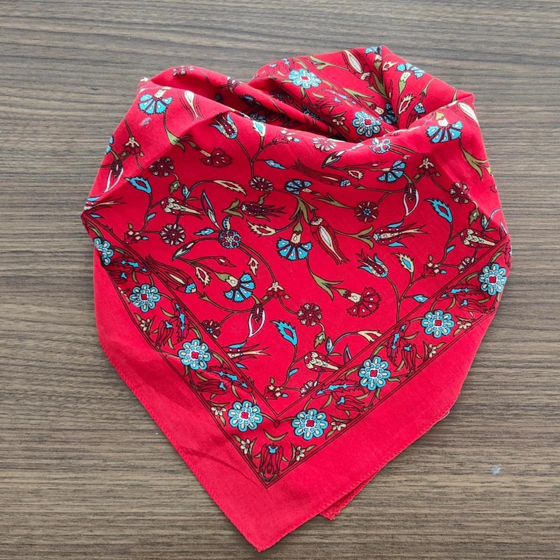 May include: A square bandana featuring a vibrant red background with a repeating floral pattern in shades of light blue, dark red, and green. The design includes stylized flowers and leaves arranged in a symmetrical layout with a decorative border.