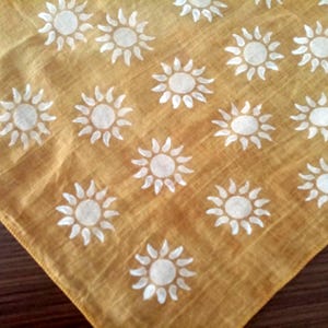 May include: Mustard yellow square fabric featuring a repeating pattern of white suns. Each sun has a central circle with radiating pointed rays. The fabric has a slightly textured appearance.