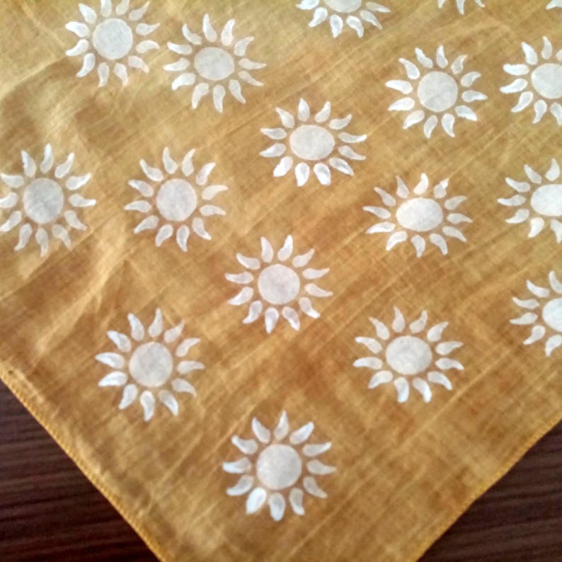 May include: Mustard yellow square fabric featuring a repeating pattern of white suns. Each sun has a central circle with radiating pointed rays. The fabric has a slightly textured appearance.