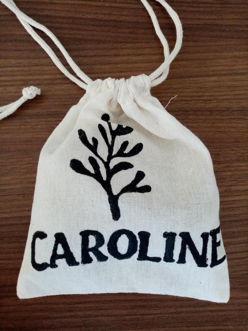 K&ouml;nnte beinhalten: Ein kleiner beigefarbener Stoffbeutel mit Kordelzug. Auf der Vorderseite befindet sich ein schwarzer Aufdruck eines stilisierten Zweiges mit Bl&auml;ttern &uuml;ber dem Namen CAROLINE in fetten, schwarzen Gro&szlig;buchstaben. Der Beutel hat zwei Kordeln.